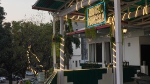 Ein Hotel mit überdachter Außenbestuhlung, einer Neonreklame und Blick auf parkende Autos und Bäume.