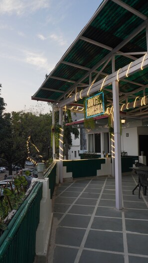 A hotel with a covered outdoor seating area, a neon sign, and a view of parked cars and trees. - Hotel Moksha (Rishikesh)