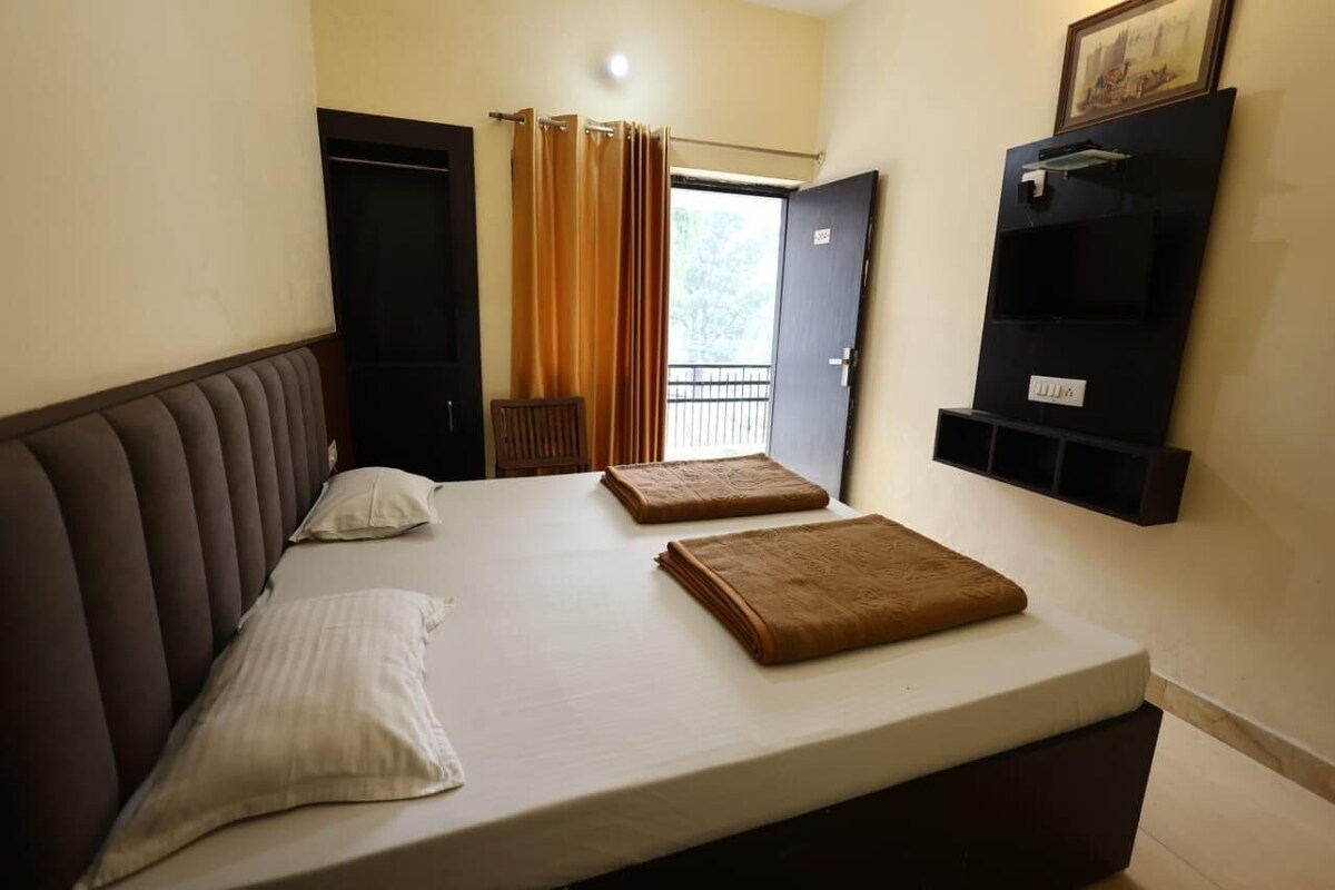 A single bed with a headboard, two pillows, and a folded blanket. There is a door with a curtain, a small shelf with a picture frame, and a window with curtains.
