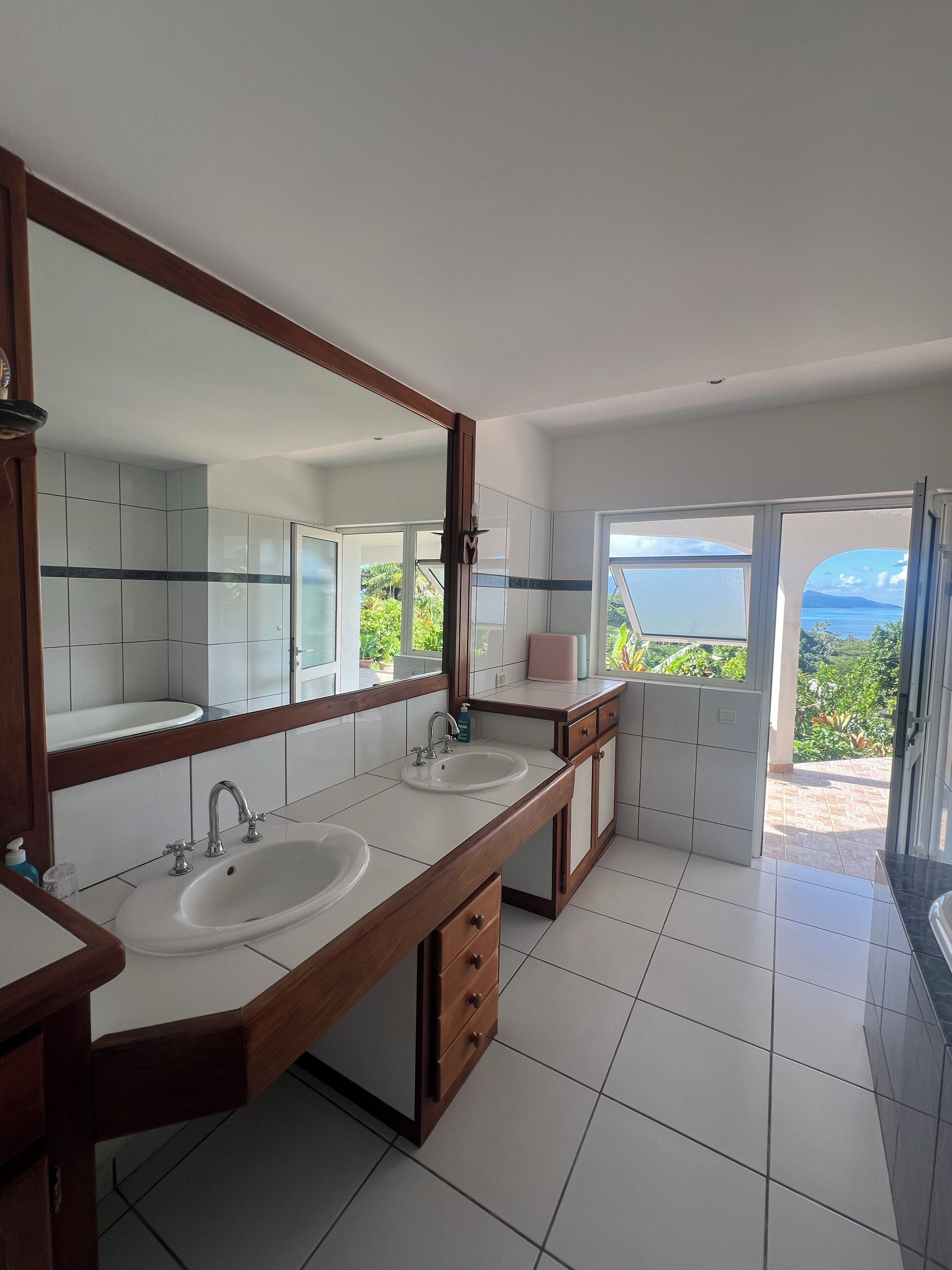 A modern bathroom with two sinks, a large mirror, and a view of a lush landscape through the door.