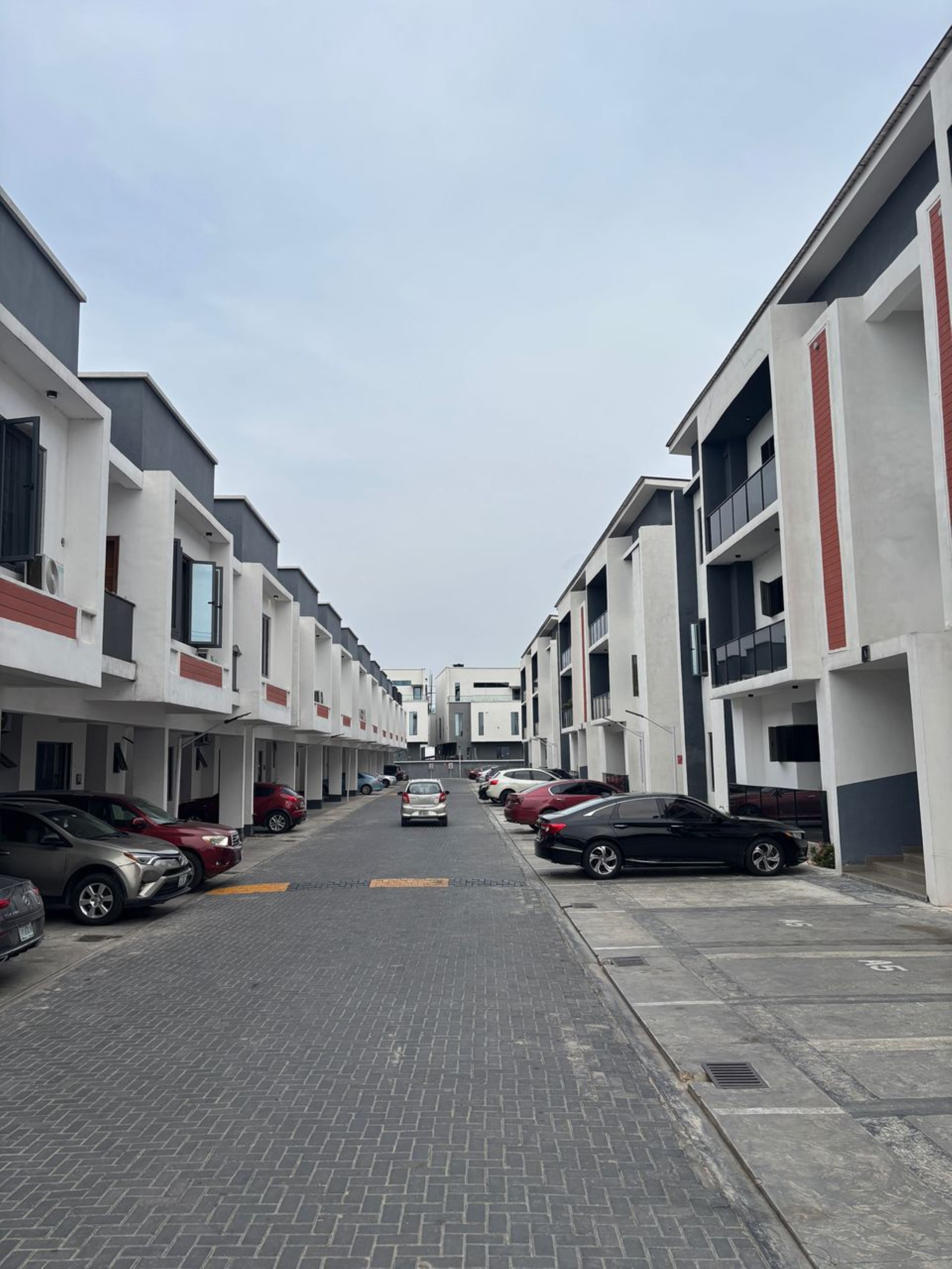 Una strada residenziale moderna con edifici a più piani, auto parcheggiate e cielo sereno.