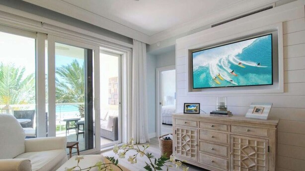 Ein modernes Wohnzimmer mit einem großen Fernseher, der eine Surfszene zeigt, einem weißen Sofa und einer Holzkombination mit Schubladen.