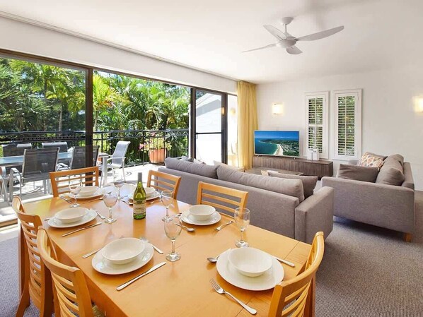 A dining area with a table set for four, a sofa, and a TV in a room with large windows and a ceiling fan.