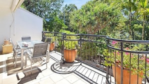 Varanda com mesa e cadeiras de vidro, rodeada por vasos de plantas e grade de metal.