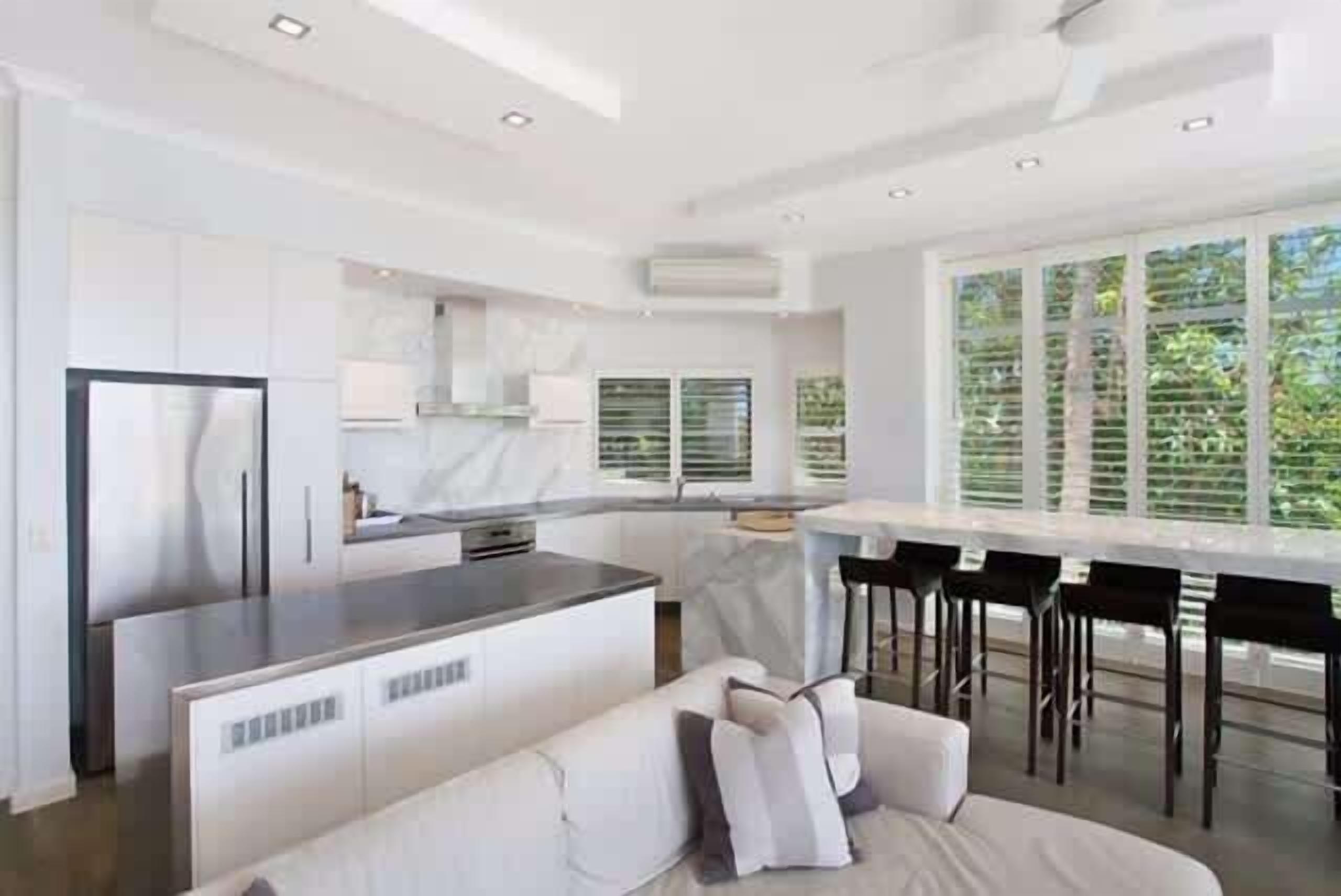 A modern kitchen with a central island, a dining area with bar stools, and a large window providing a view of greenery.