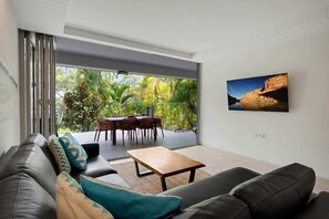 Un salon moderne comprenant un canapé, une table basse et un coin repas avec des chaises et une table. Une grande fenêtre offre une vue sur la verdure environnante.
