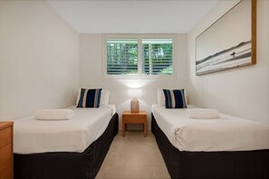 A hotel room with two beds, a nightstand with a lamp, a window with blinds, and a painting on the wall. - Little Cove Beach 3 Bedroom Unit 01 (Noosa Heads)