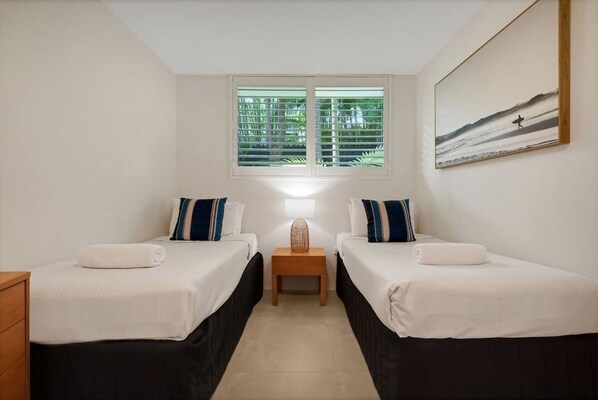 Une chambre d’hôtel avec deux lits, une table de chevet avec une lampe, une fenêtre avec des stores et un tableau au mur.