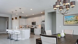 A modern kitchen with white cabinets, stainless steel appliances, a central island with bar stools, and a dining area with a dark wood table and chairs. - Impeccable 3 BR / 2 BA home with expansive lake view - Serenity Garden (Venice)