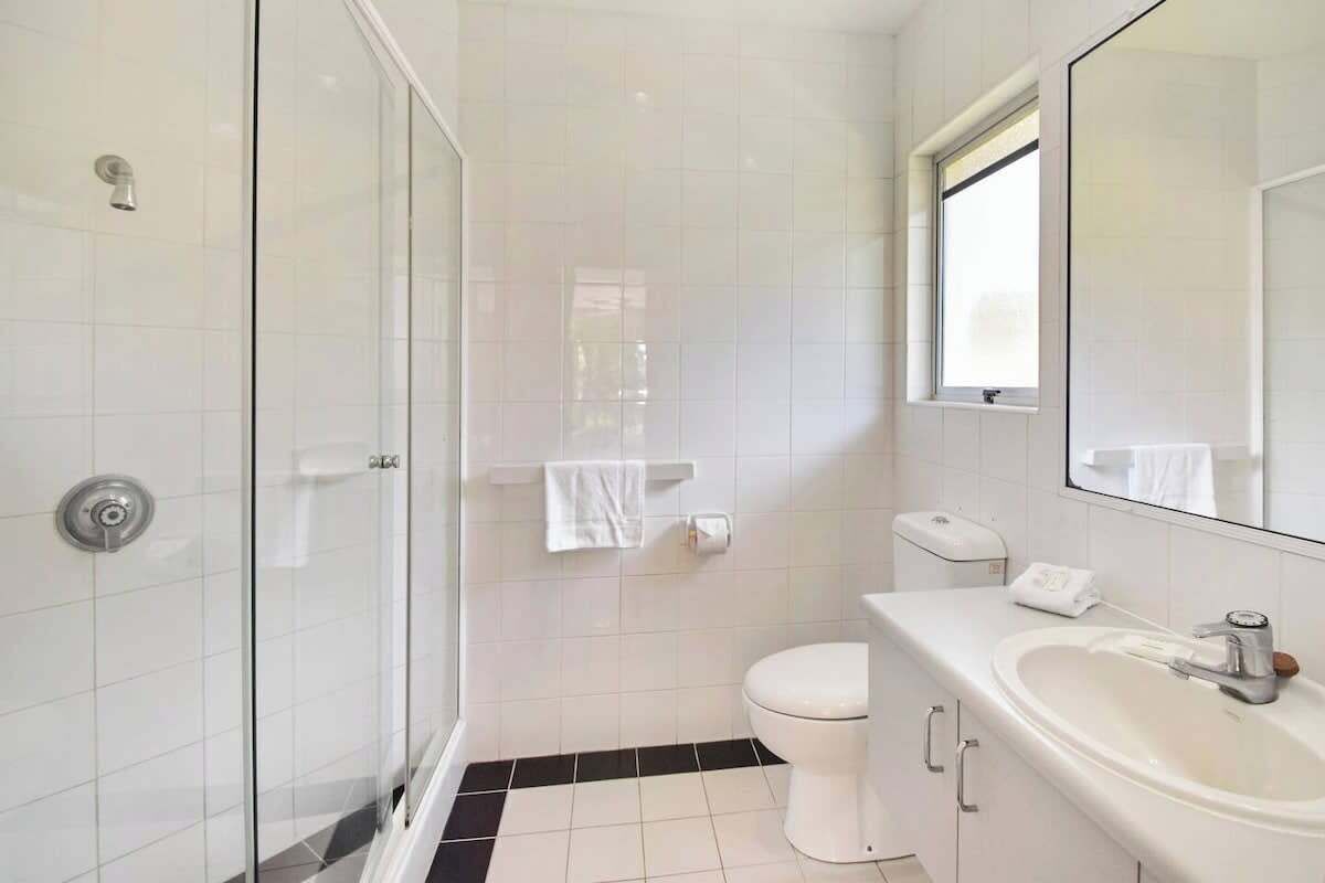 A bathroom with a glass shower enclosure, a white toilet, a sink with a mirror, and a window.