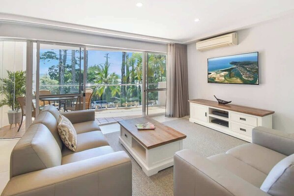 A modern living room with a large sliding glass door leading to a balcony, a flat-screen TV mounted on the wall, and a wooden coffee table.