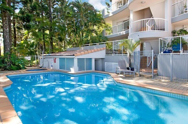 A swimming pool with a sun deck, surrounded by trees and a multi-story building with balconies.