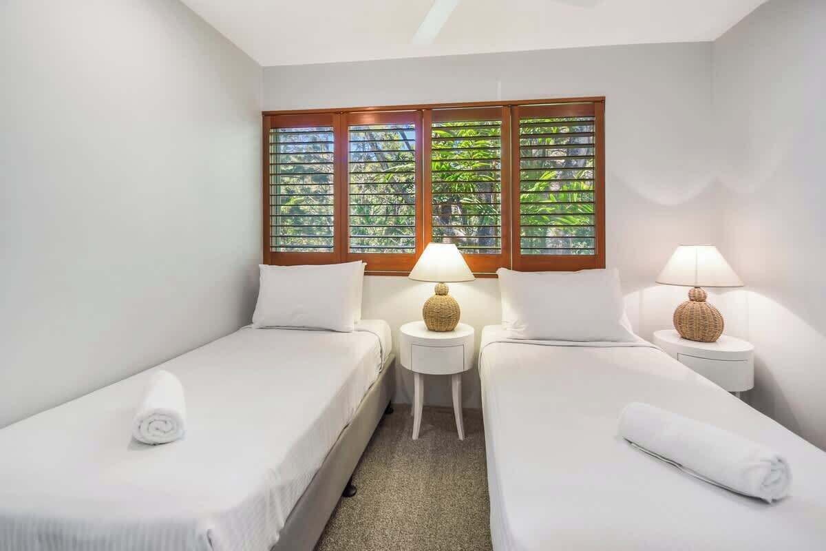 Ein Hotelzimmer mit zwei Einzelbetten, einem kleinen Tisch, zwei Lampen und einem Fenster mit Blick auf Grünflächen.