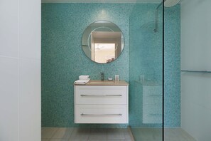 Casa de banho com parede revestida de azulejos, espelho redondo, lavatório com produtos de higiene e zona de duche.