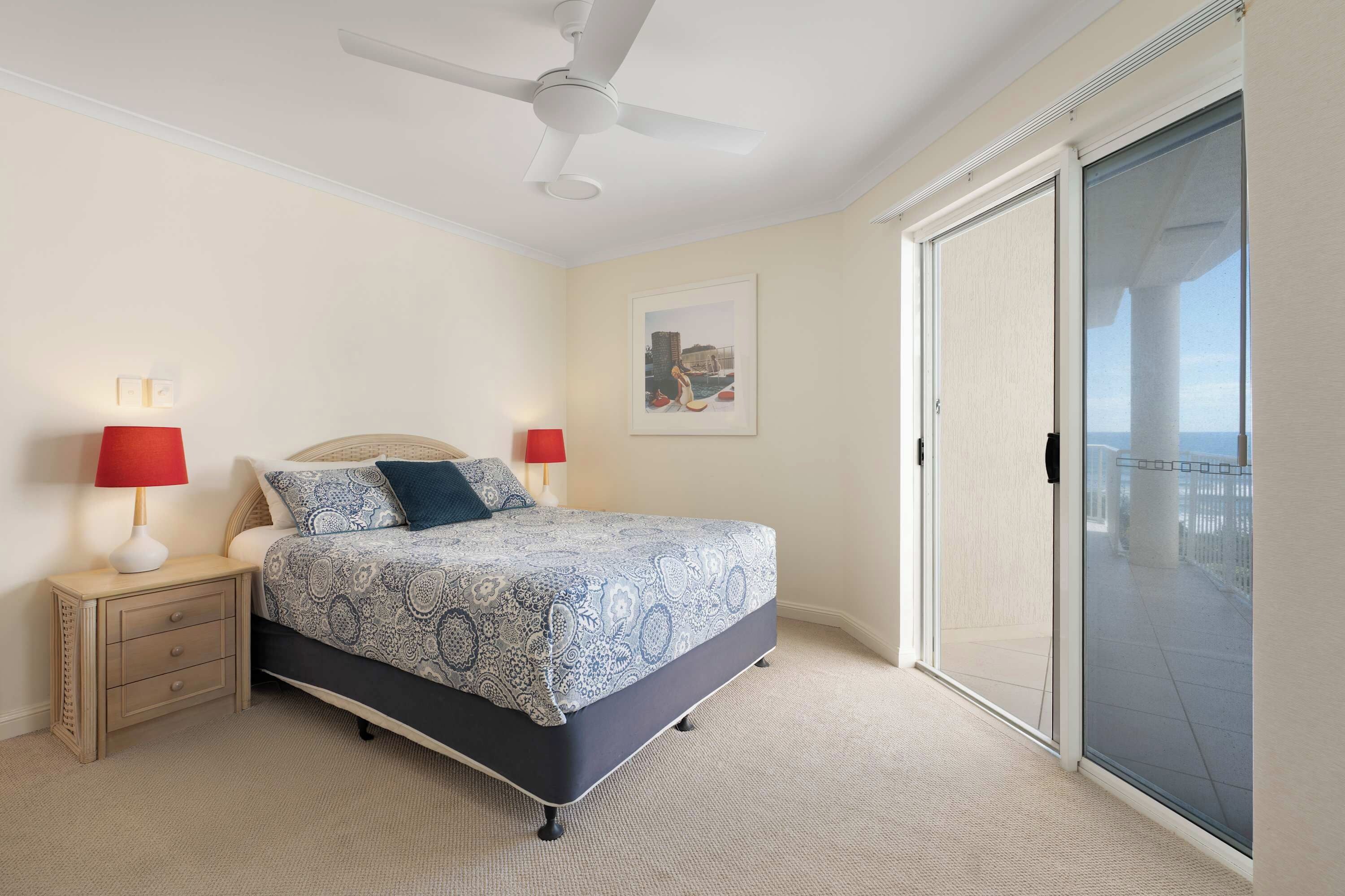 A bedroom with a bed, two bedside tables, a ceiling fan, a large window with a view, and a door.