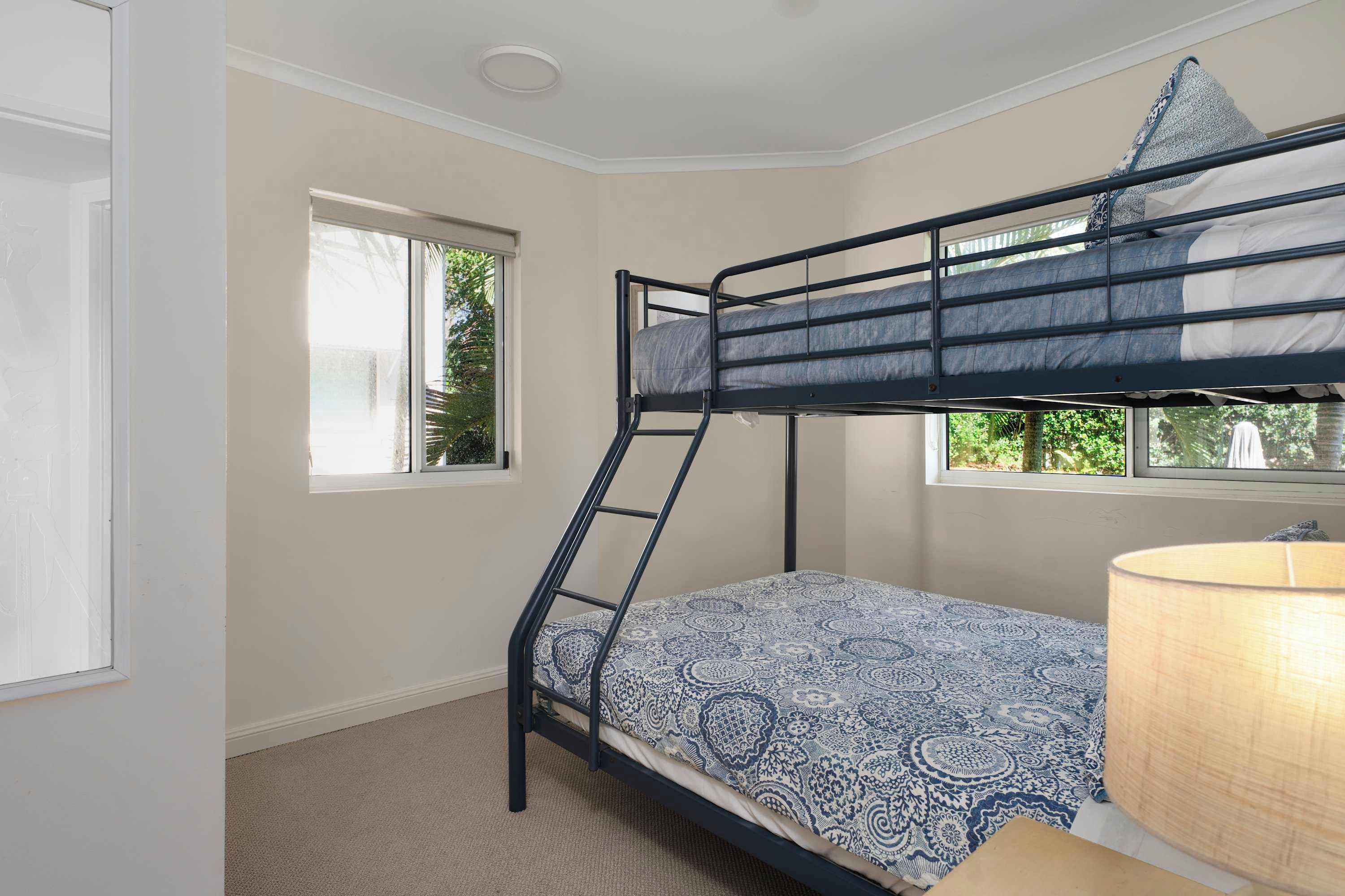 A bunk bed with a patterned bedspread, a window with a view of greenery, and a lamp on the floor.
