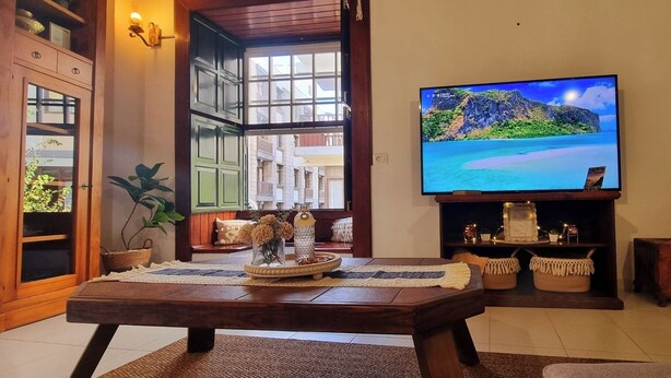 Uma sala de estar com uma mesa de centro de madeira, uma TV exibindo uma paisagem de praia tropical e uma lareira com objetos decorativos.