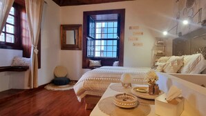 Um quarto aconchegante com uma cama grande, uma mesa de cabeceira com objetos decorativos, uma janela com cortinas e um quadro na parede com fotografias emolduradas.