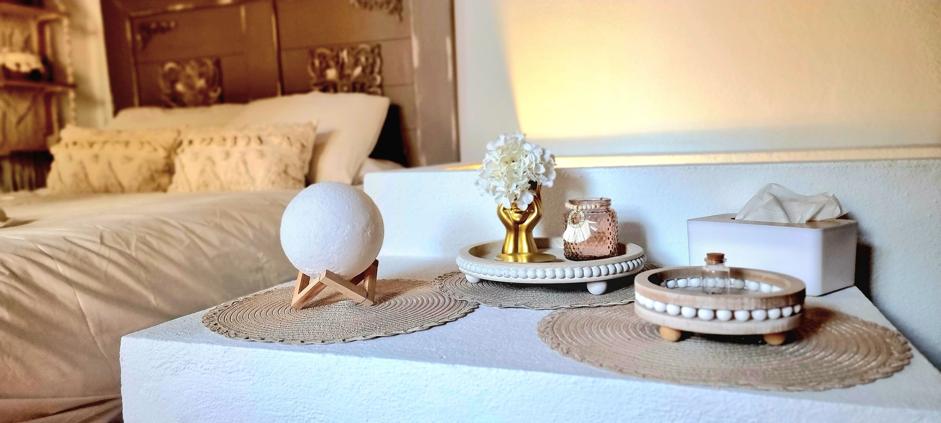 Quarto bem arrumado com cama, mesa de cabeceira e objetos decorativos, incluindo um vaso com flores, um pote e uma caixa de lenços.