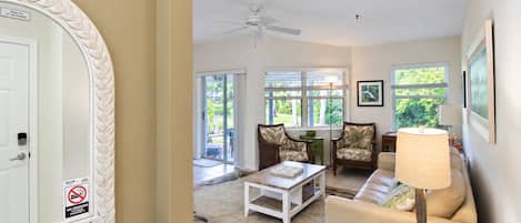 A living room with a ceiling fan, a sofa, a coffee table, and a lamp. There is a door with a 'No Smoking' sign.