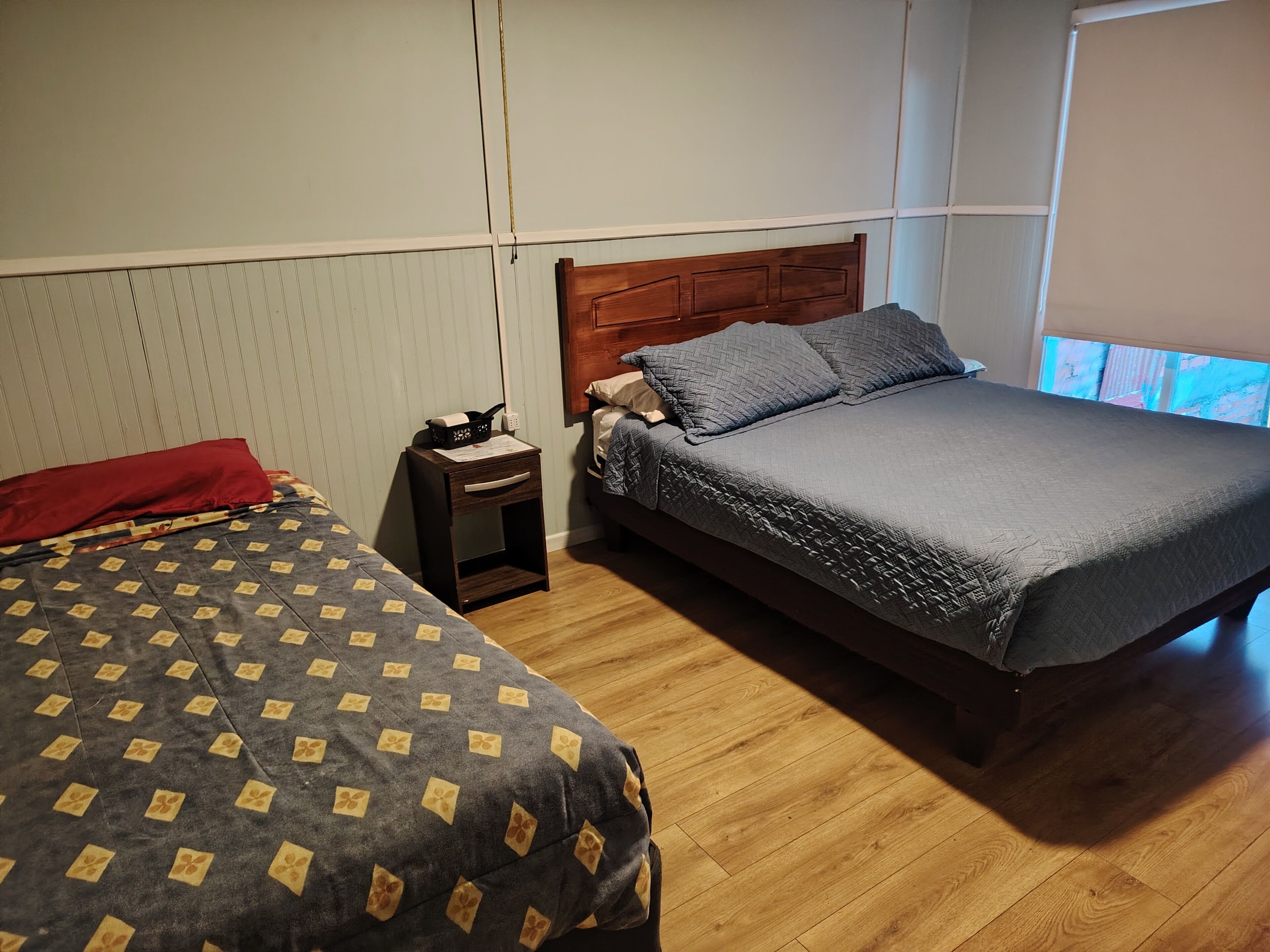 A bedroom with two beds, a wooden headboard, a nightstand, and a window with blinds.