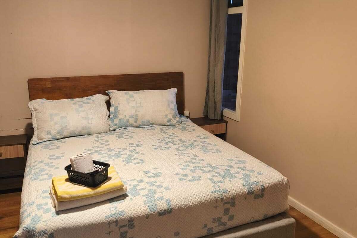Un lit simple avec une tête de lit en bois, une table de chevet, une commode et une fenêtre avec des rideaux.
