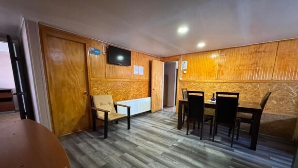 A room with a wooden wall panel, a television mounted on the wall, a round table with chairs, and a wooden floor.