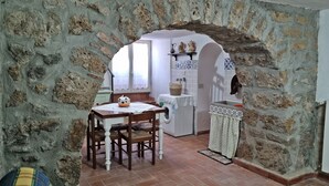 A rustic room with a stone wall, a dining table with chairs, a washing machine, and a small kitchen area with a sink and decorative items. - Nives Holiday House, home in the ancient castle walls in the historic center. (Tuscania)