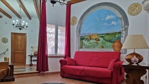 A living room with a red sofa, a glass coffee table, a wooden door, a chandelier, and a mural on the wall. - Nives Holiday House, home in the ancient castle walls in the historic center. (Tuscania)