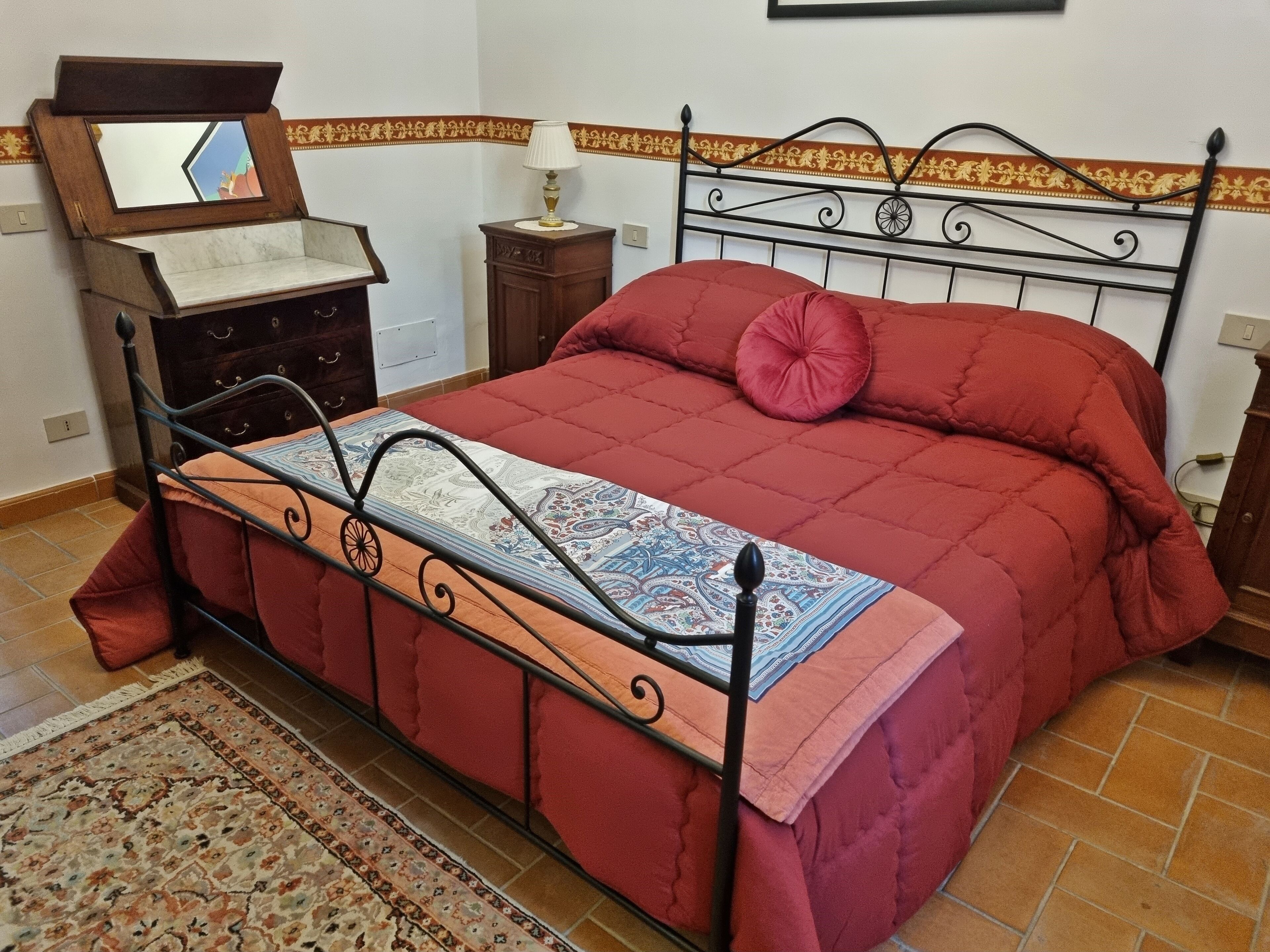 Un lit avec un cadre en métal, une couette rouge et une couvre-lit à motifs. Une petite table de chevet en bois avec une lampe se trouve à côté du lit. Un miroir décoratif est accroché au mur au-dessus du lit.