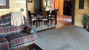A cozy living room with a plaid sofa, a coffee table with a book and plants, a dining area with a wooden table and chairs, and a kitchen visible through an open doorway. - Lake cabin 2-bedroom sleeps 6.  1/2 mile from trails.  Backus with private dock. (Backus)