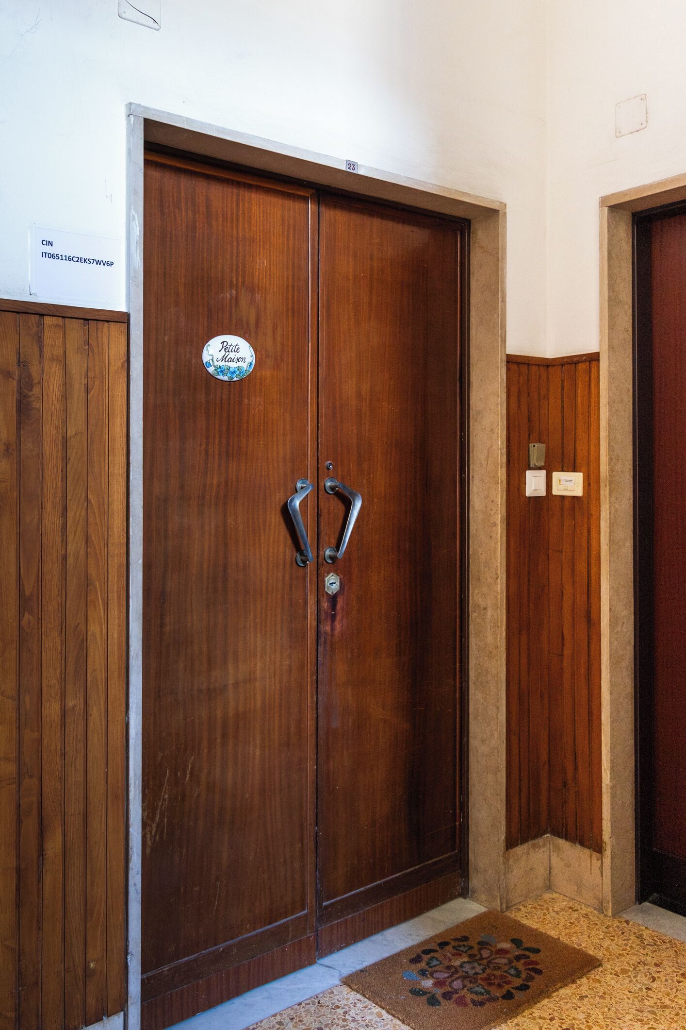 A wooden double door with a sign above it and a doormat in front.