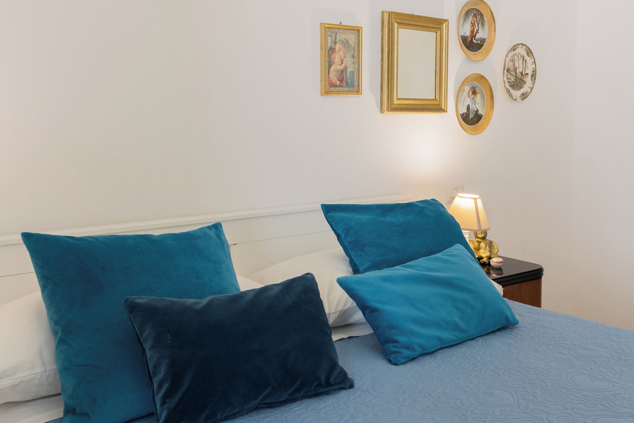 Una cama con almohadas color turquesa y azul oscuro, una lámpara de noche y decoración en la pared, incluyendo cuadros y un plato.