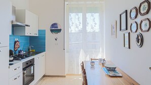 Une cuisine avec des meubles blancs, un fourneau et des carreaux bleus. On y trouve une table à manger avec des chaises et une fenêtre avec des rideaux.
