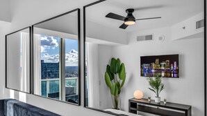 Une chambre moderne dotée d’une grande fenêtre, d’un ventilateur de plafond, d’une vue sur la ville, d’un téléviseur à écran plat et d’un canapé bleu foncé.