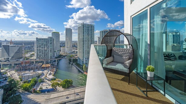 Un balcon situé dans un immeuble de grande hauteur, avec un fauteuil en osier, une vue sur une rivière et des bâtiments modernes en arrière-plan.