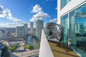 A high-rise balcony with a wicker chair, a view of a river, and modern buildings in the background.