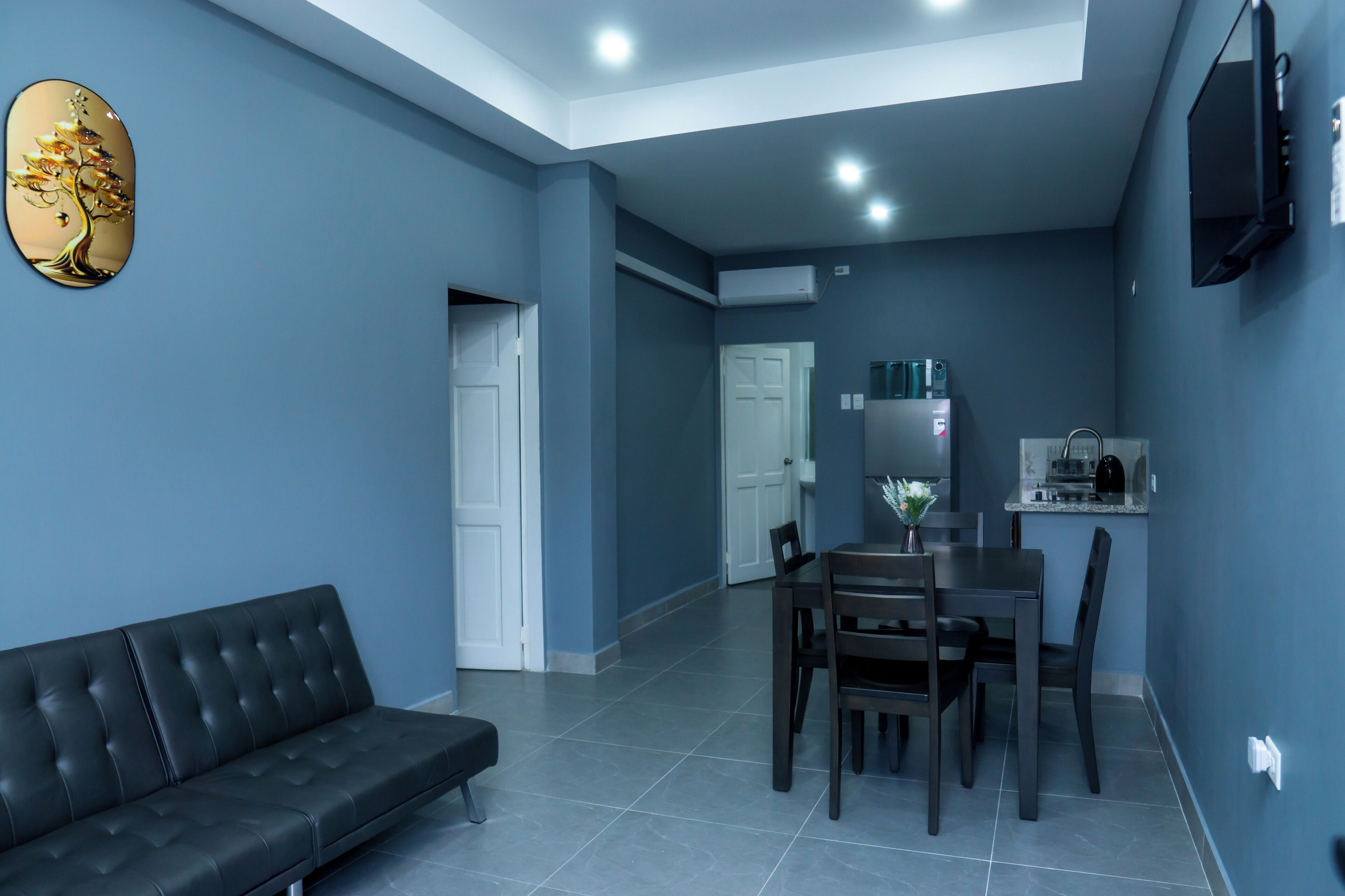 A modern living room with a black leather sofa, a dining table with chairs, and a kitchen area with a refrigerator and microwave.