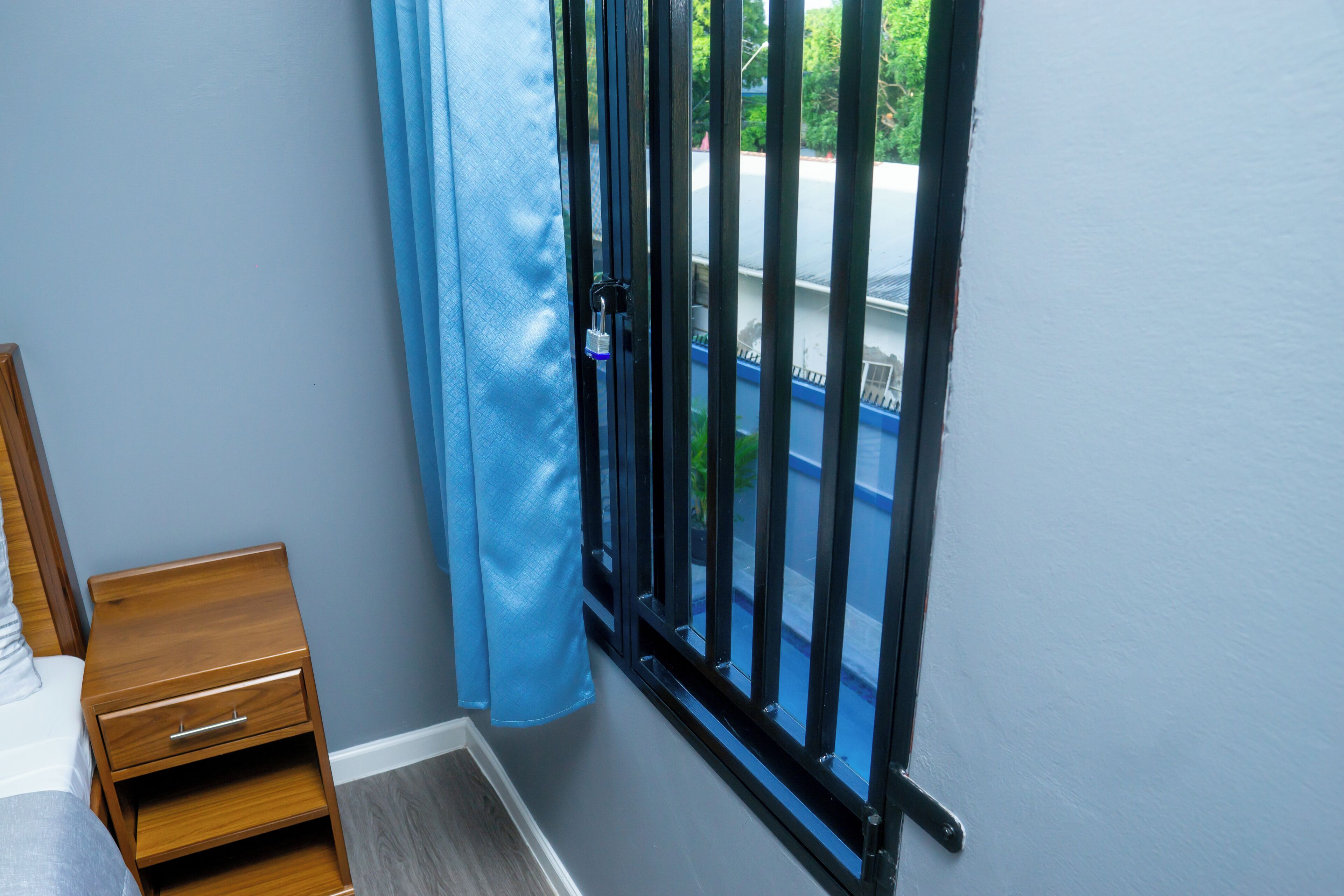 Une pièce avec une table de chevet en bois, un rideau bleu et une vue sur une rue, visible à travers un portail en métal noir.