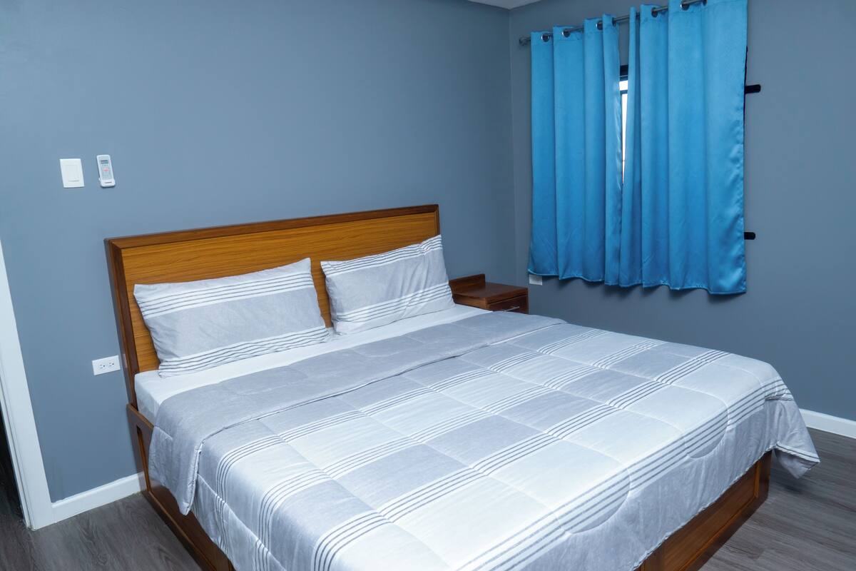 A neatly made bed with a wooden headboard, a nightstand, and a blue curtain.