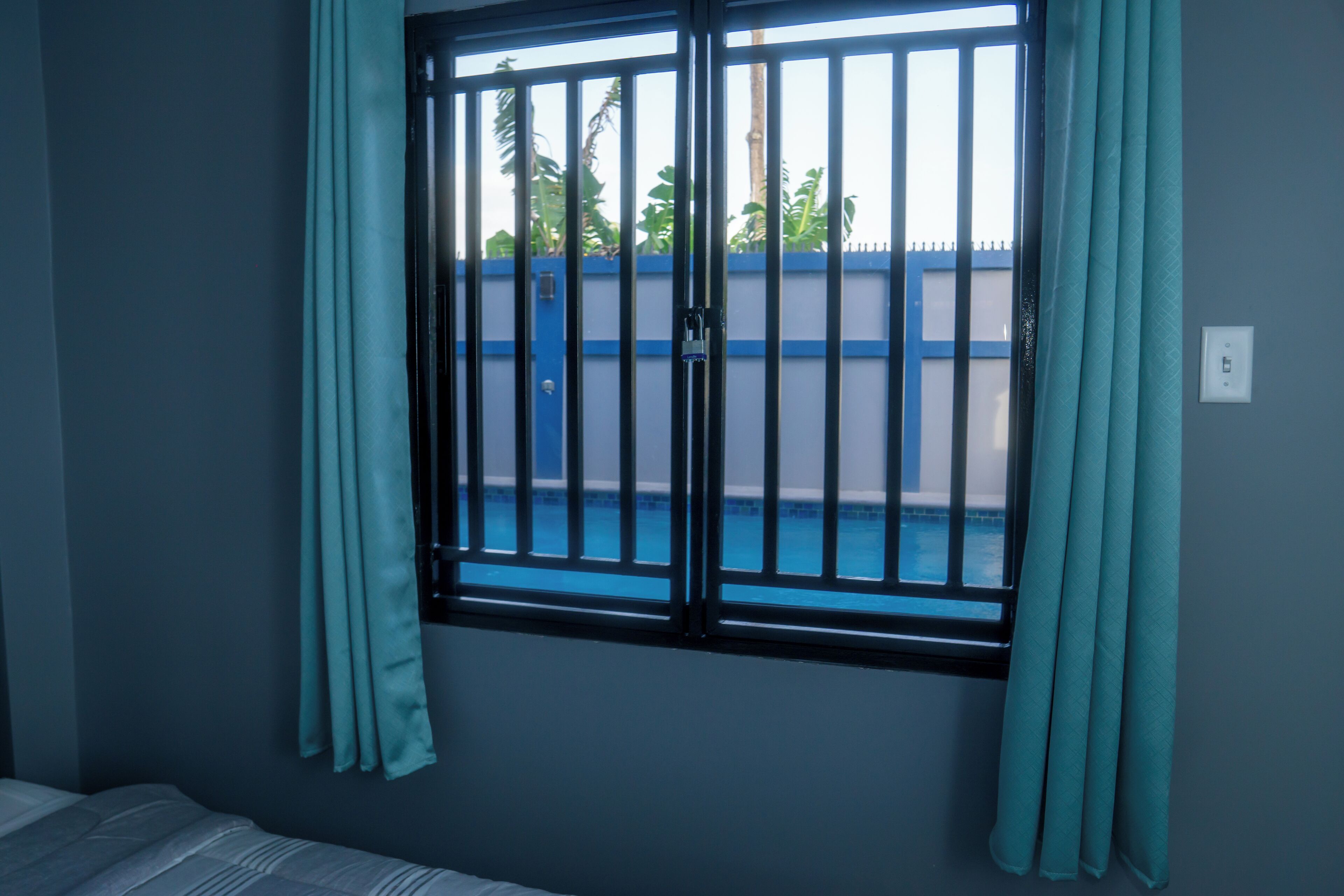 Ein Zimmer mit einem Fenster, das ein Metallgitter und türkisblaue Vorhänge zeigt. Es gibt ein Bett und einen Blick auf eine blaue Wand und Grünpflanzen draußen.