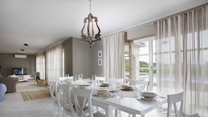 A dining area with a white table set for four, surrounded by white chairs, a chandelier, and large windows with sheer curtains. - Villa Sara Bajkini with vineyard view (Bajkini)