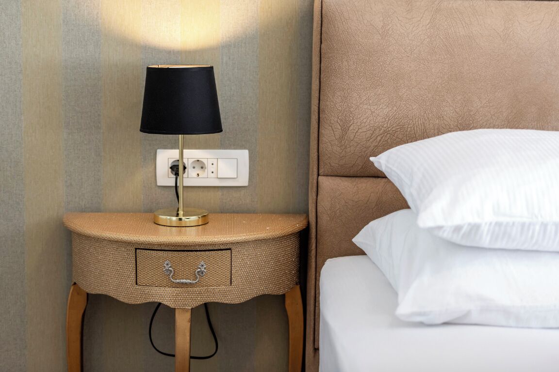 Un lit bien fait, avec du linge de lit blanc et une tête de lit, une lampe de chevet avec un abat-jour noir et une table de chevet en osier dotée d’un tiroir.