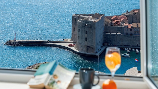 Une vue d’une ville côtière, avec un château imposant, une jetée et un verre de jus d’orange posé sur une table.