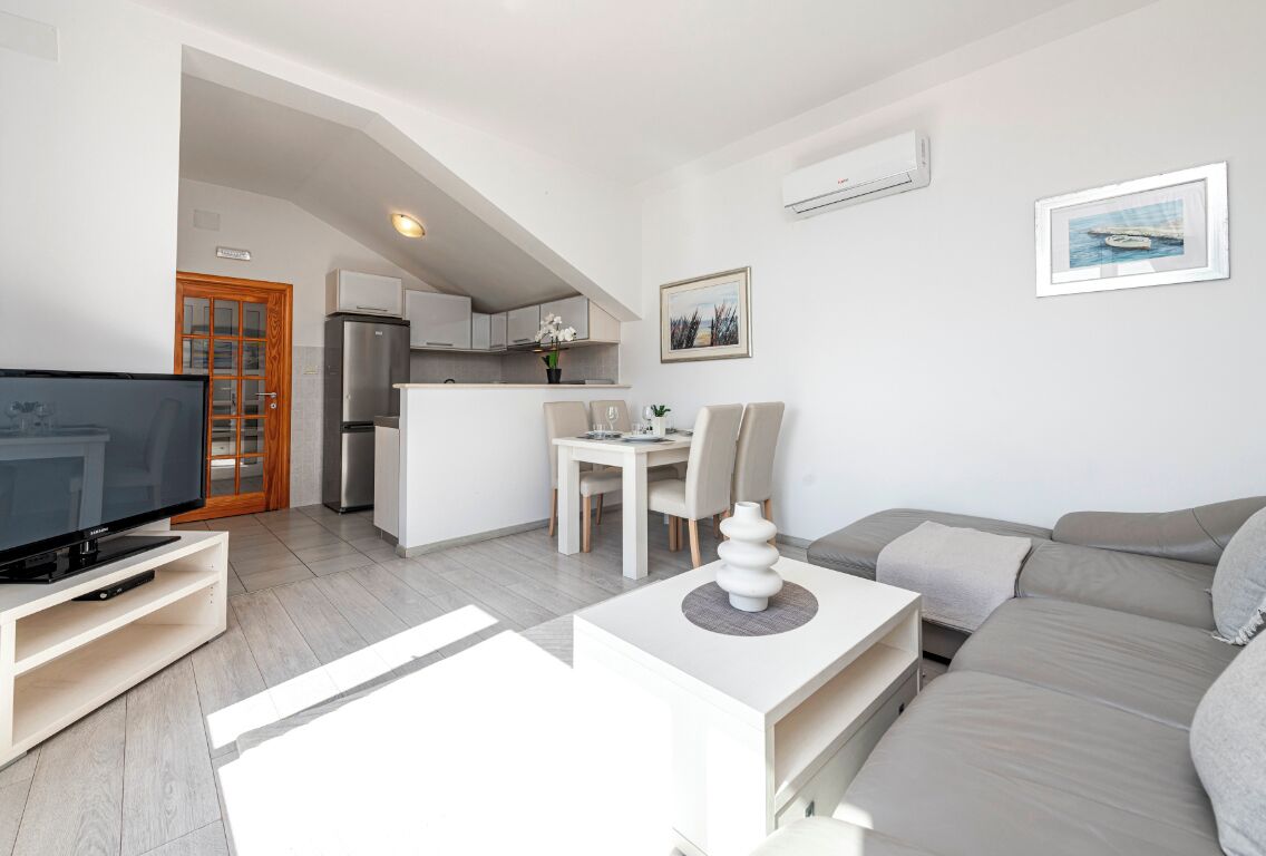 A modern living room with a flat-screen TV, a sofa, a dining table with chairs, and a kitchen area in the background.