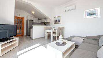A modern living room with a flat-screen TV, a sofa, a dining table with chairs, and a kitchen area in the background.