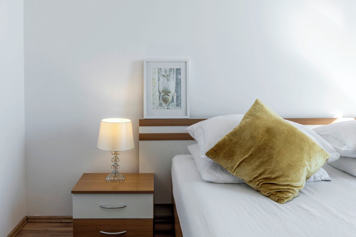 Una cama bien tendida con sábanas blancas y una única almohada color mostaza, una lámpara de noche sobre una mesita de madera y una obra de arte enmarcada en la pared.