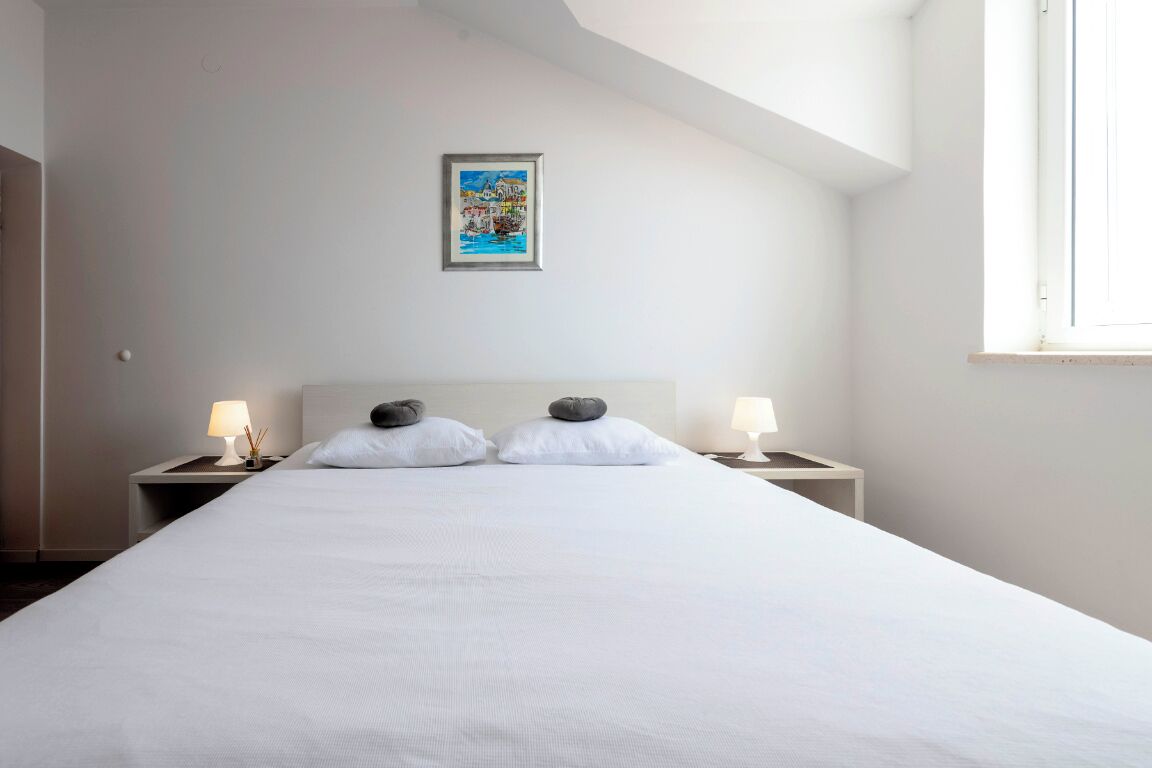 Ein ordentlich bezogenes Bett mit weißen Leinentüchern und zwei Kissen, daneben Nachttische mit Lampen, in einem Zimmer mit schrägen Wänden und einem einzelnen Fenster.