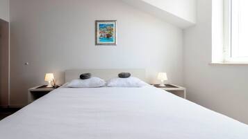 Ein ordentlich bezogenes Bett mit weißen Leinentüchern und zwei Kissen, daneben Nachttische mit Lampen, in einem Zimmer mit schrägen Wänden und einem einzelnen Fenster.