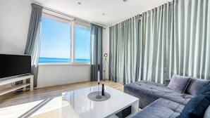 Un salon moderne doté d’une grande fenêtre, d’un téléviseur à écran plat, d’un canapé gris et d’une table basse blanche.